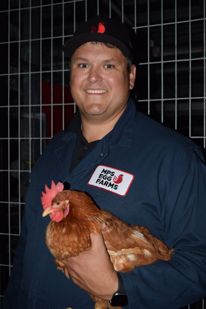 Un hombre, TJ Klein, sostiene un pollo rojo dentro de una instalación avícola. 