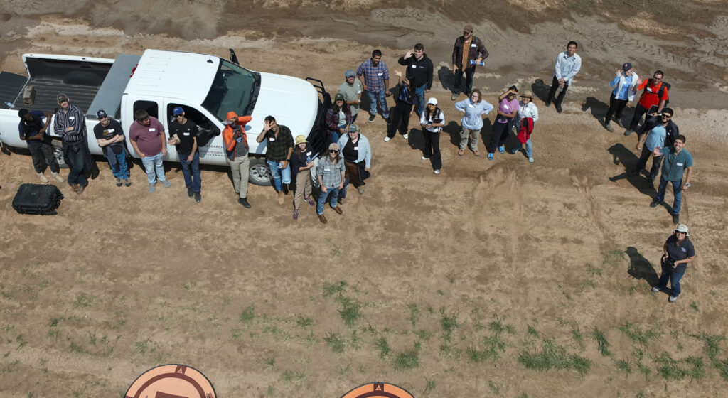 about 25 students and instructors stare up into the sky to look at a drone that is taking their picture in a field
