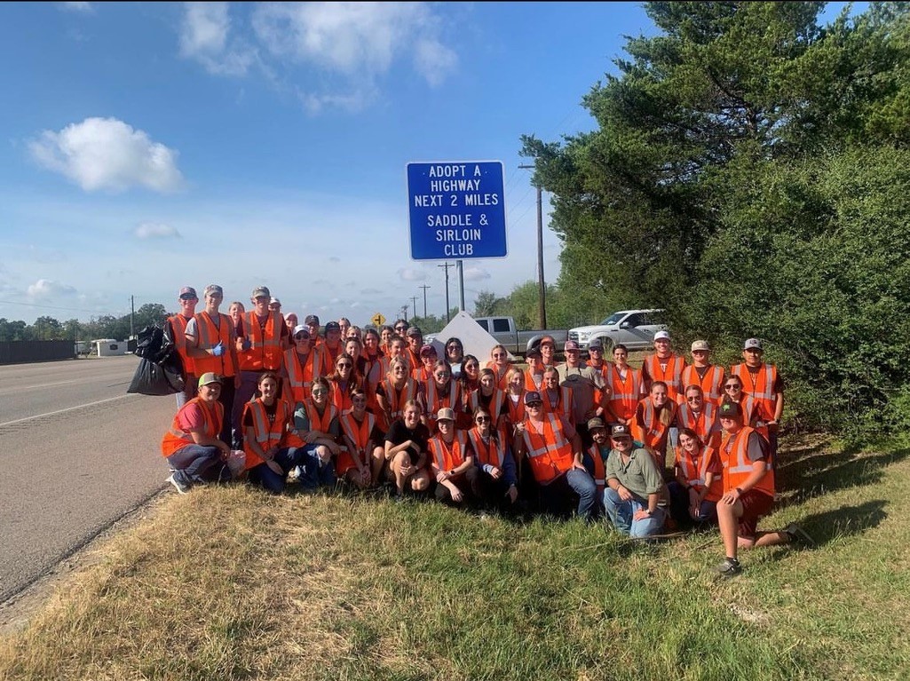 A group of students wearing orange vests pose together on the side of the road with a sign that reads: Adopt a highway next 2 miles Saddle & Sirloin Club.