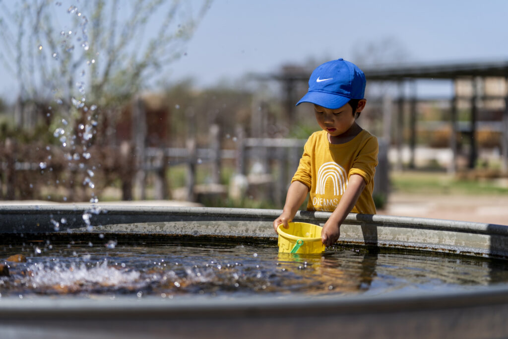 Young child with blue cap standing in the sun and dipping a yellow plastic bucket into a water tank