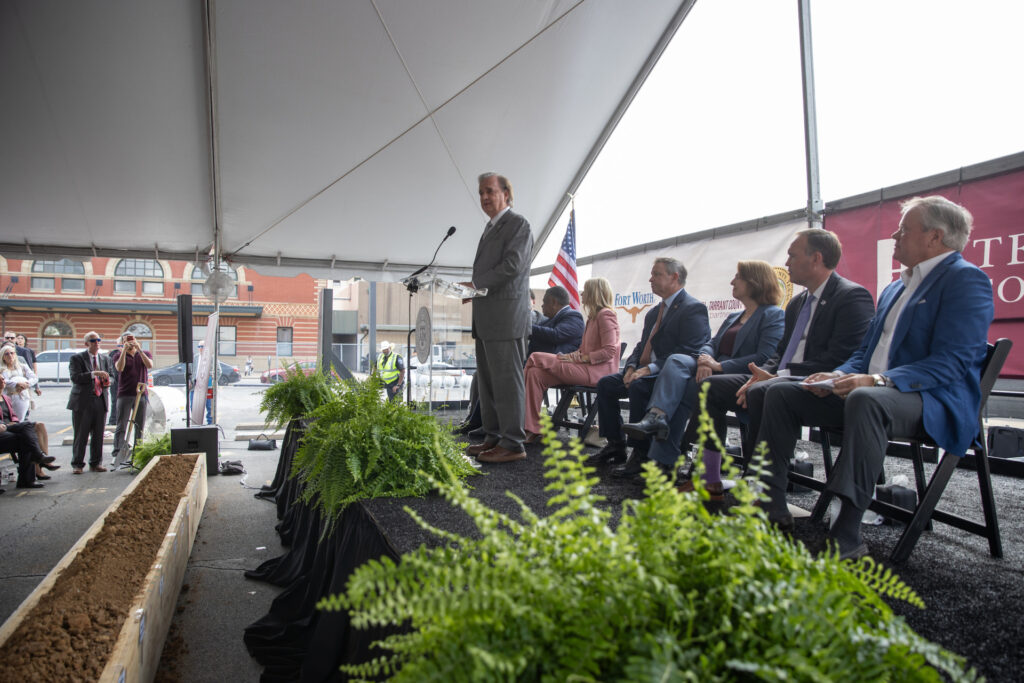 Announcing the new Texas A&M-Fort Worth campus, a man stands at a podium on a plant-lined stage with people seated behind him and others can be seen in front