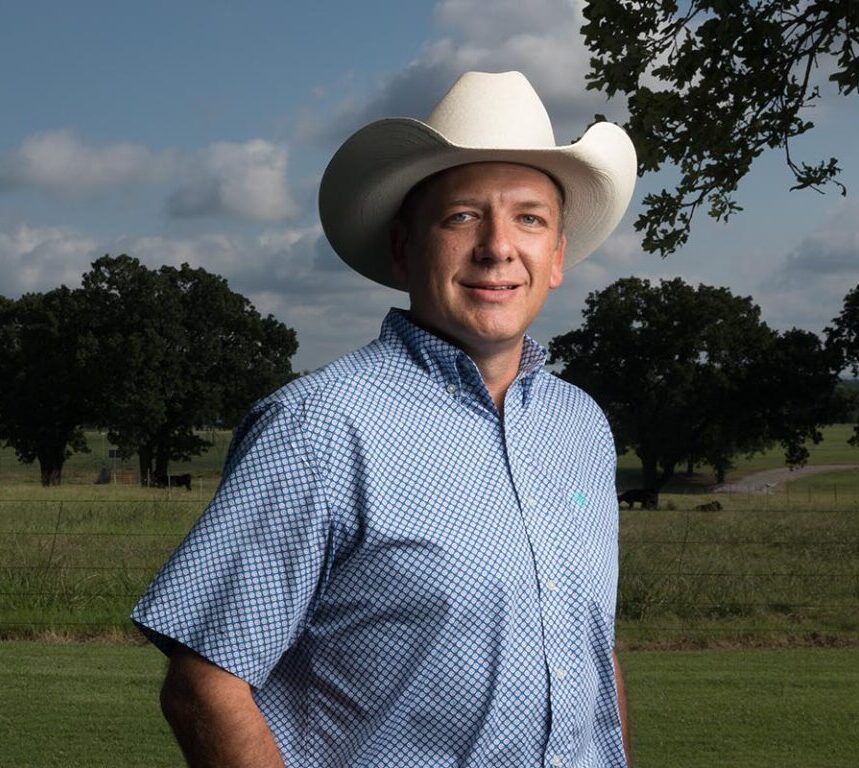 Un hombre que lleva un sombrero de vaquero se para en un campo con árboles detrás de él