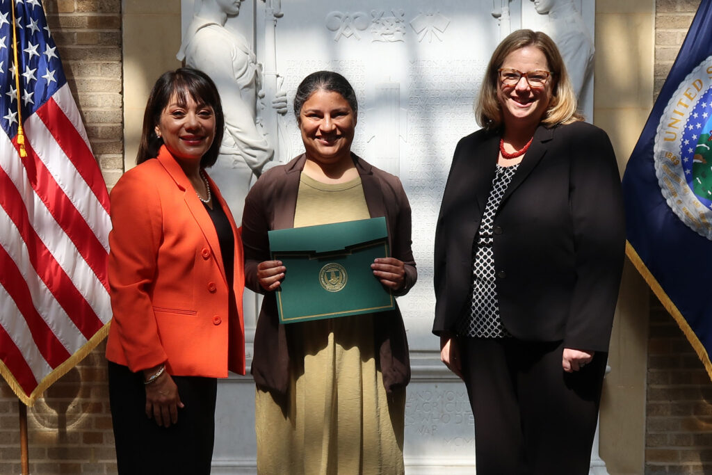 Three women stand between the U.S. flag and the USDA flag. The middle one holds a green folder with an E. Kika De La Garza Fellow certificate in it