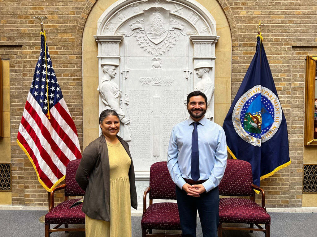 a woman and a man stand between two flags in the USDA building while participating in the E. Kika De La Garza Fellows Program
