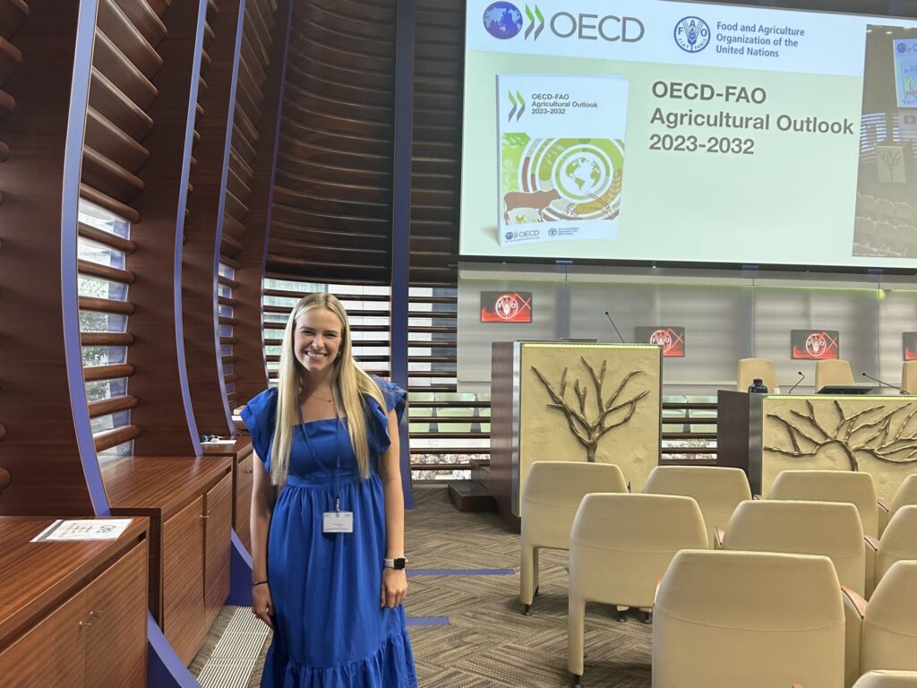 Katelyn Klawisnky standing in a room wearing a blue dress with a projector screen behind her that reads "OECD-FAO Agricultural Outlook 2023-2032".