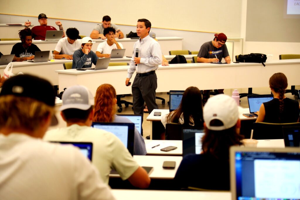 Nick Kilmer standing in the middle of a lecture hall holding a microphone while lecturing to students, many of whom have laptops in front of them