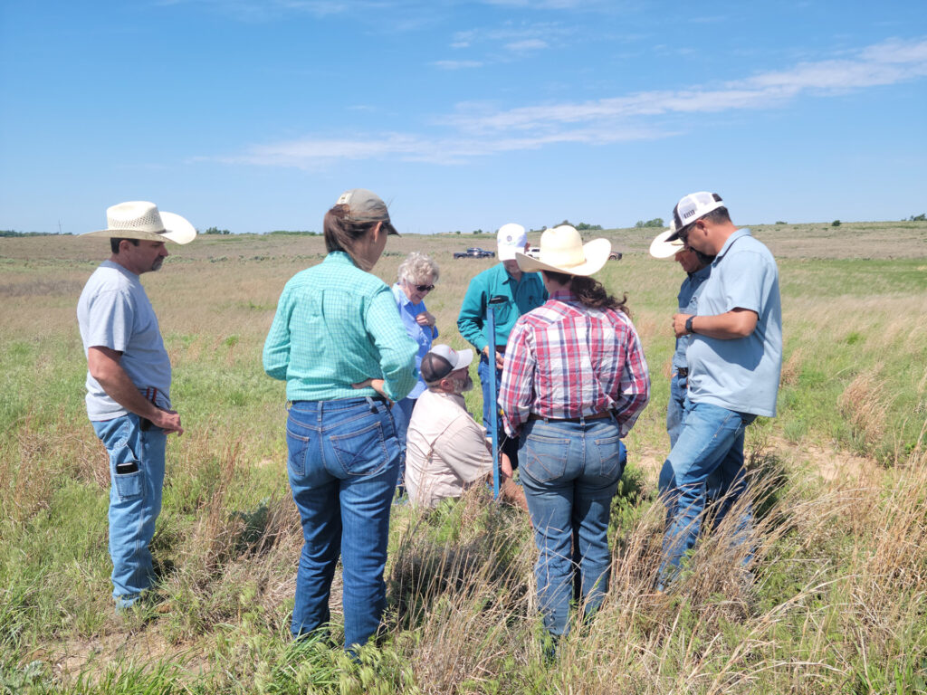a man kneels down in the center of a circle of men and women ranchers standing in a ranch pasture