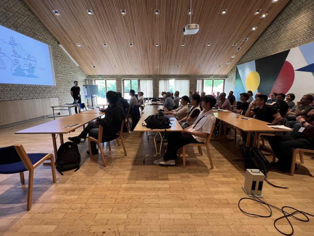 A presenter projects image slides onto a wall for researchers gathered in a large hall with a sloped, modern wood-panel ceiling, modern wood furniture and brick walls. Natural colors and large modern abstract painting covering one wall.