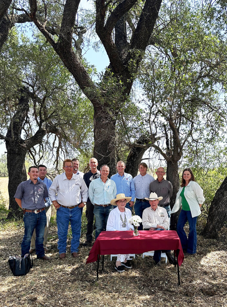 A man and a woman sitting at a table with maroon table cloth and a vase of flowers center piece. A group of 10 people stand behind them. They are in a group at a ranch with trees in the background and hay-scattered on the foreground on the Slaughter Ranch.