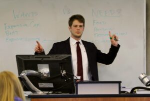 Photo of a man in a coat and tie in front of a whiteboard with a computer in front of him while he teaches a class