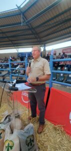 A man in a khaki button-up short sleeve shirt at a podium holding and speaking into a microphone. Standing on straw floor with a steel fence behind him and an audience sitting behind the fence.