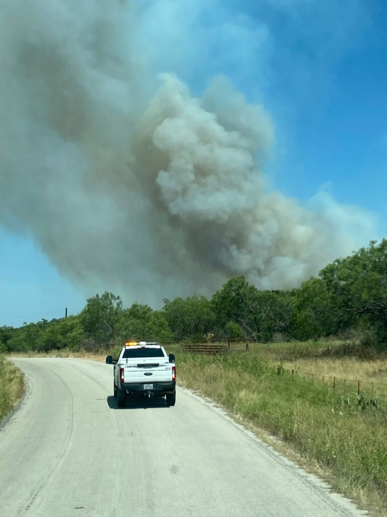 a large plume of gray smoke rises into a blue sky above green trees as a pickup drives down the road warning of wildfire danger