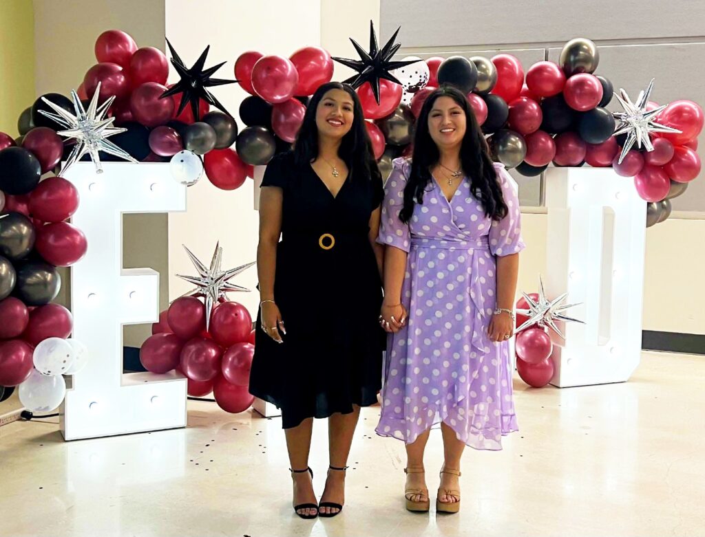 Two women standing in front oof block letter decorated with red, silver and black balloons. 