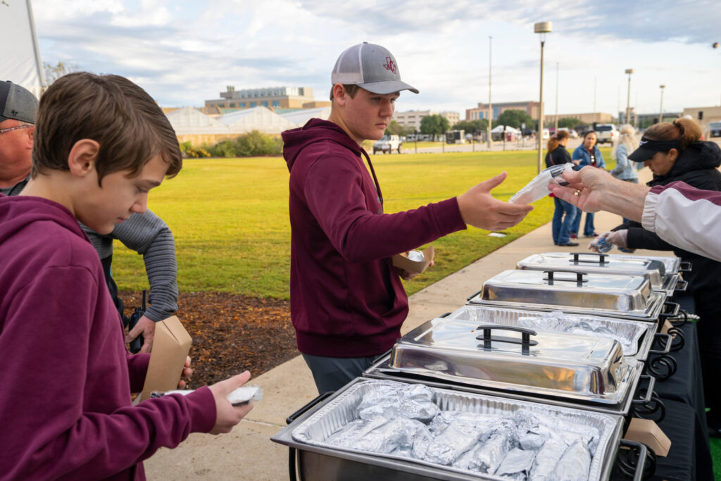 A Texas A&M Aggie Tailgate where teenagers wearing maroon sweatshirts are in line to eat. Food is in steel serving containers with warming units under them to maintain a safe temperature.