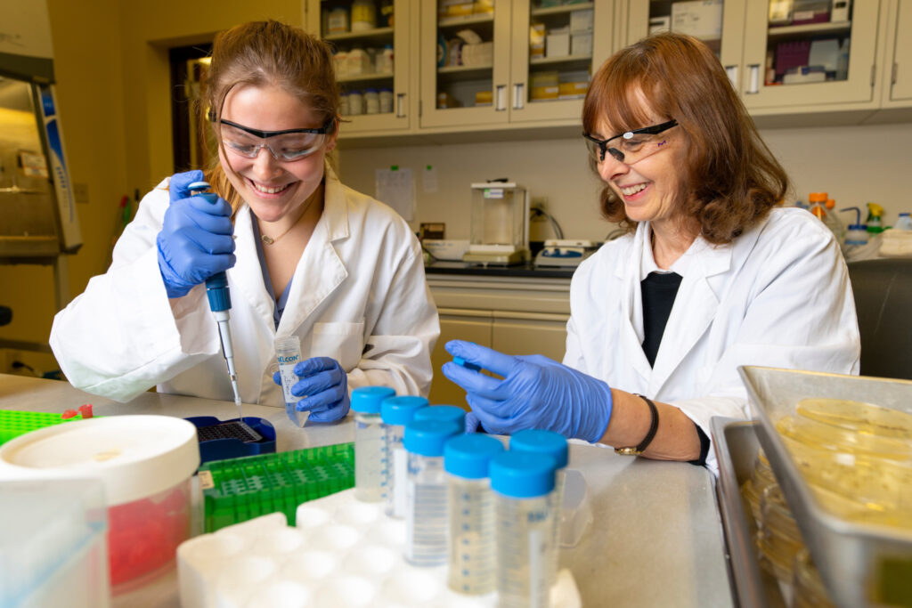 two ladies working in a lab wearing lab coats, rubber gloves and protective glasses. 