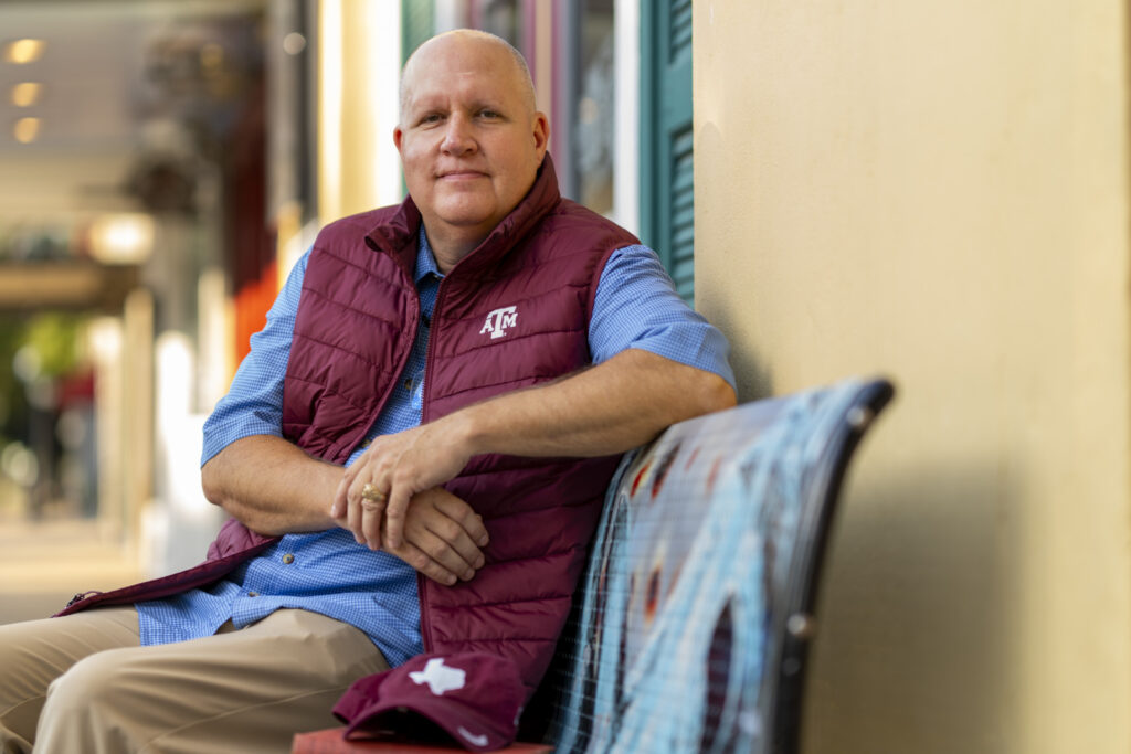 A man sitting on a bench wearing khakis, a blue short and a maroon sleeveless jacket with the Texas A&M logo on it. 
