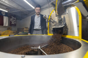 A man wearing a blue blazer stands watches as coffee beans are dumped form a roaster.