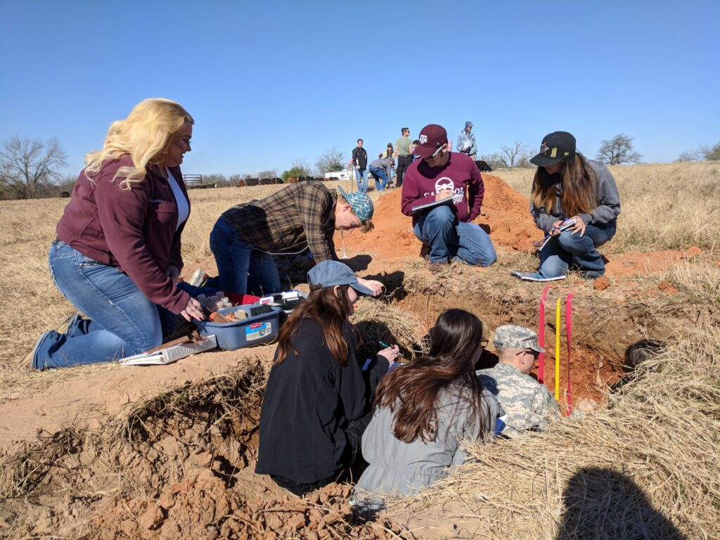 Group of male and female college students taking soil samples in a trench in a field.