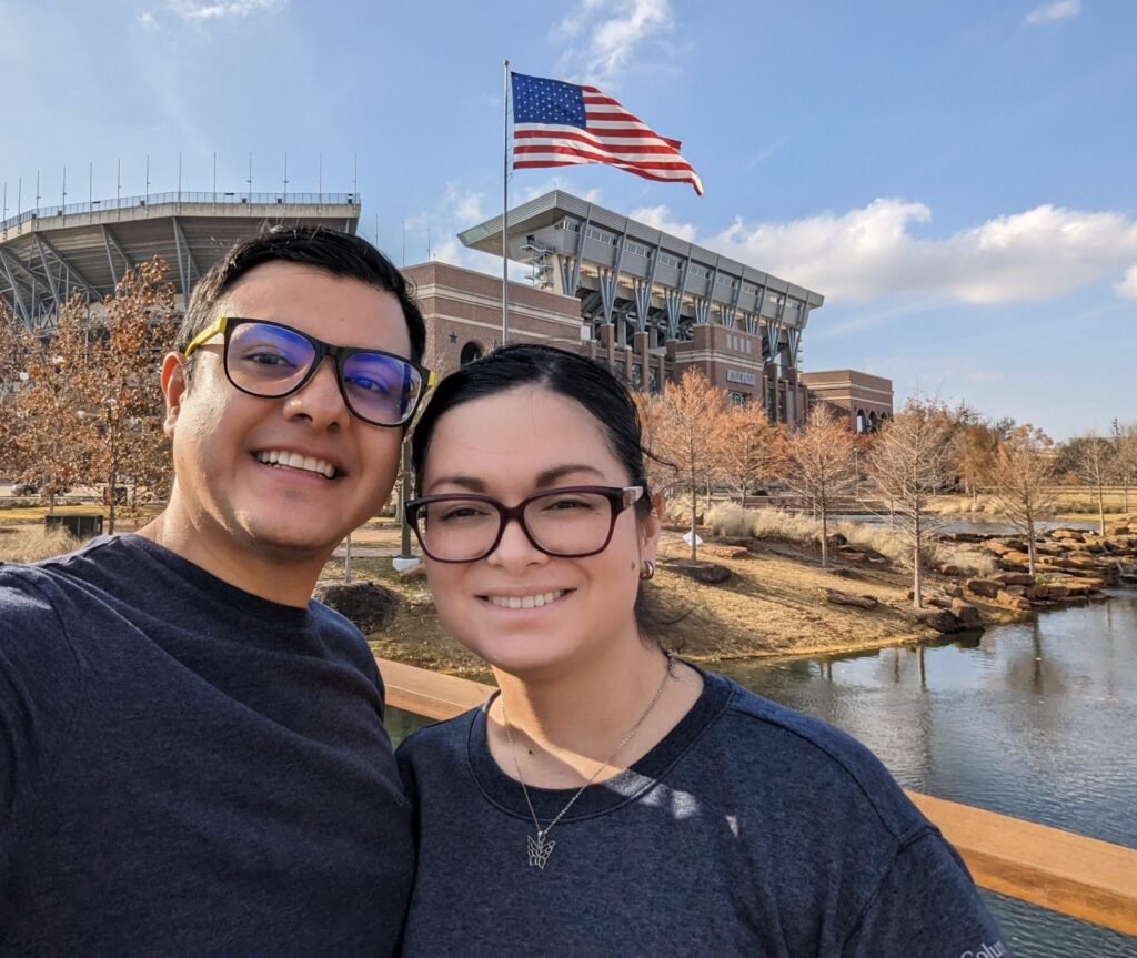Doctoral canddidate Karla Solis Salazar smiles next to her spouse outside a large stadium with an American flag and a river in the background
