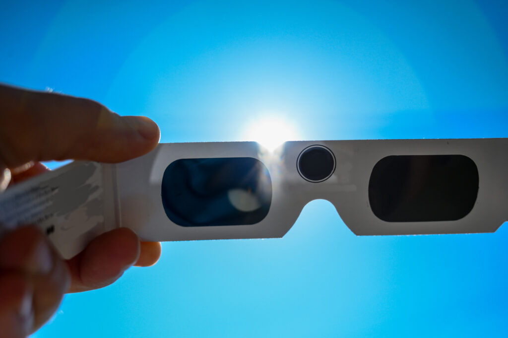 a hand holding eclipse viewing glasses with the sun in the background. 