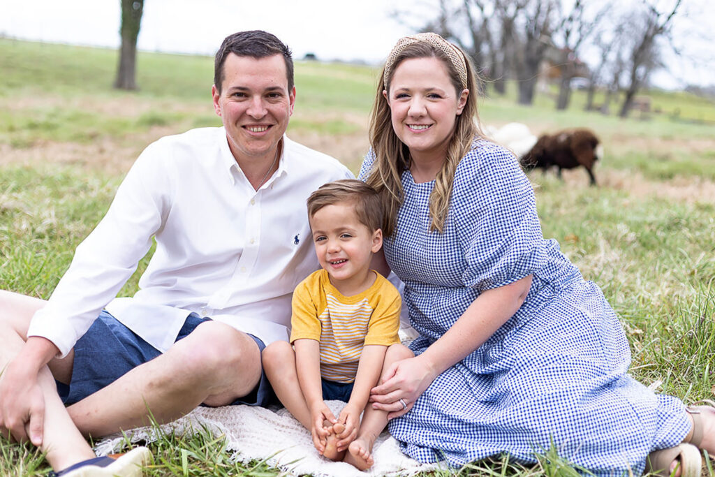 A young man and woman sit in a grass field with their son between them. Gatlan and Rachel Gray and Brigham.