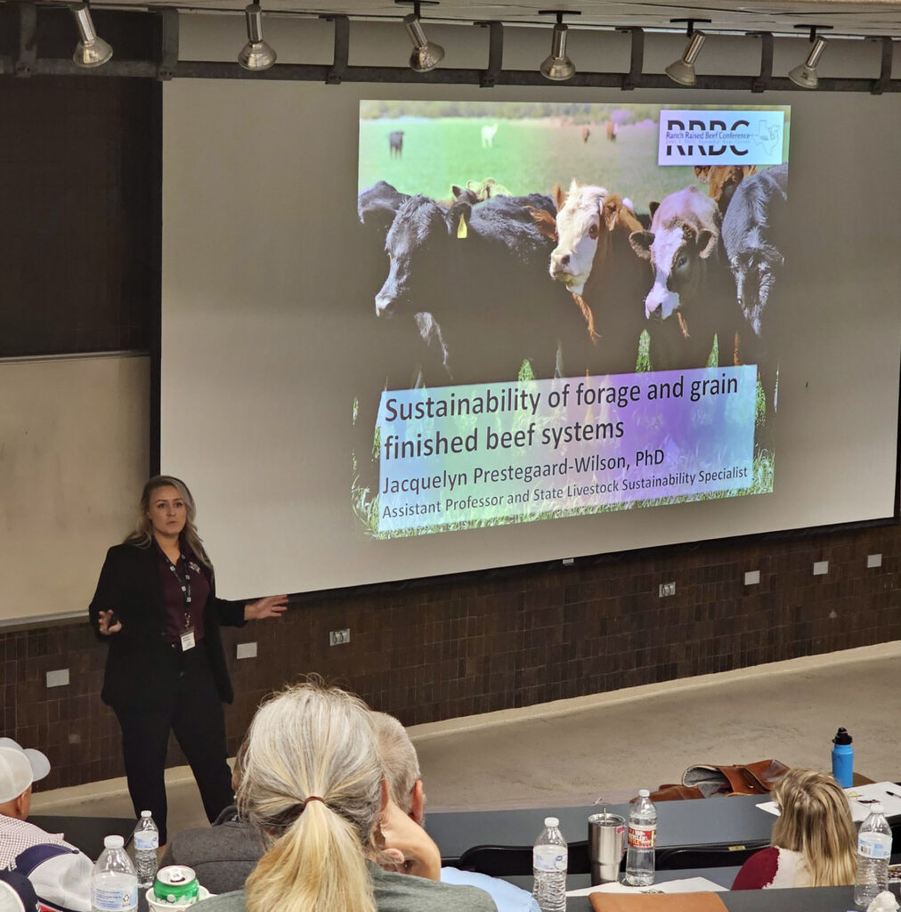 a woman stands at the front of the room presenting on sustainable livestock systems. The slide says Sustainability of forage and grain finished beef systems Jacquelyn Prestegaard-Wilson, PhD