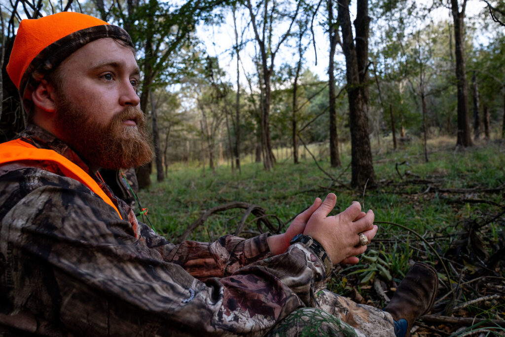A man in camouflage and hunter orange sits and waits for deer. 