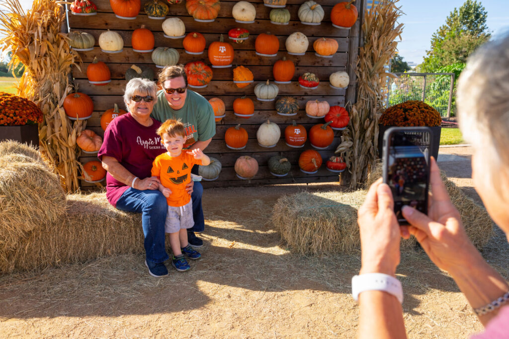 Older woman, young mother and toddler pose for a photo at the pumpkin wall at the Fall Festival in The Gardens at Texas A&M University.