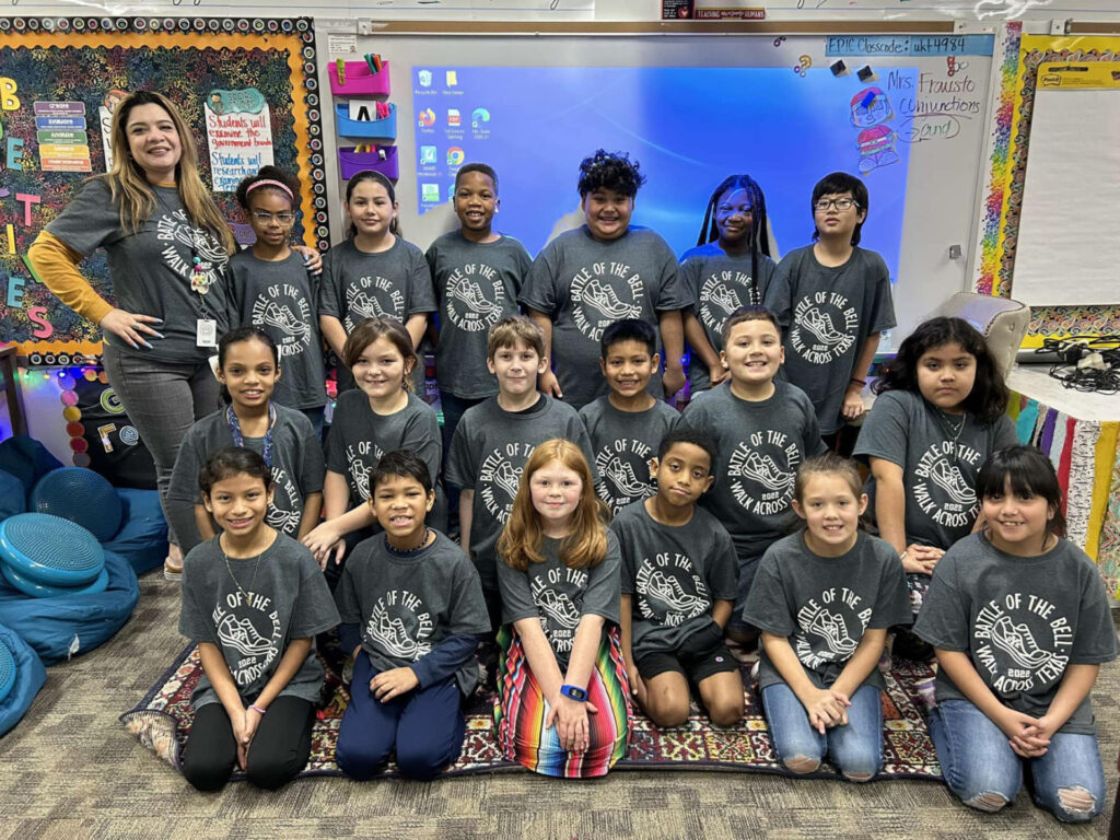 Students kneeling and standing together in their classroom wearing grey Walk Across Texas T-shirts. 