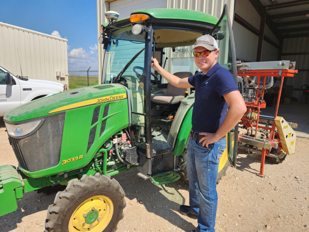 a man, Brandon Gerrish, stands holding on to a John Deere green tractor with a planter/cultivator behind it. Gerrish is the new AgriLife Extension small grains specialist