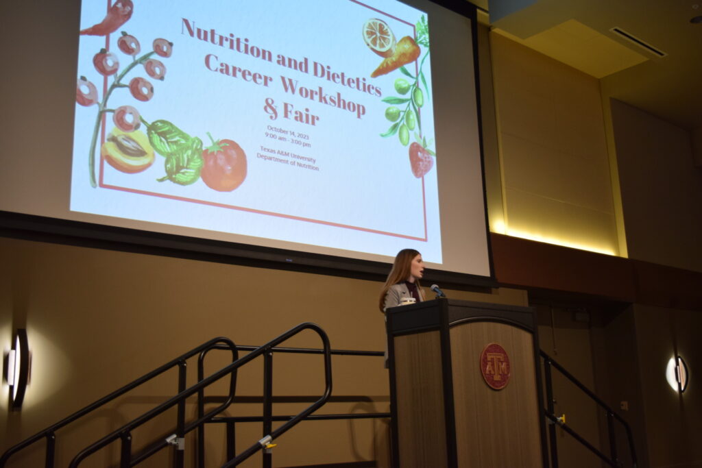 A woman stands behind a lectern while speaking. The screen behind her has fruits and vegetables on it and has the words Nutrition and Dietetics Career Workshop and Fair. 
