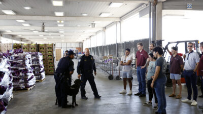 Two officers in uniform with show K-9 Rottweiler to a group of people in street clothes. The group is in a large warehouse with rows of garage doors and bags and boxes of goods stacked about 8 feet high.