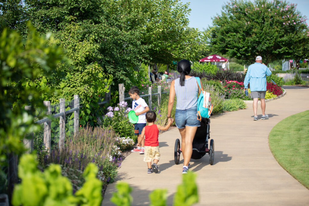 A woman pushes a stroller with two young children walking along a path at The Gardens at Texas A&M University. 
