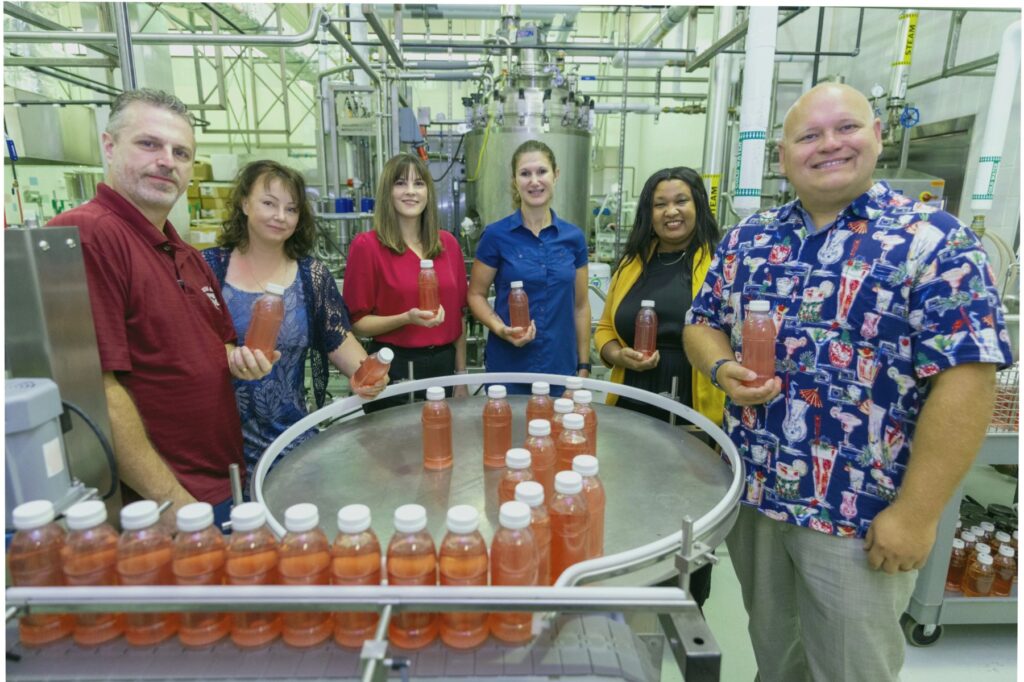 Group photo of four women and two men standing around a conveyor belt holding bottles of juice that are being used in their project focusing on chronic disease prevention