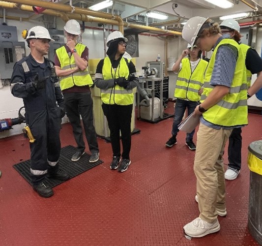 A group of four people in hardhats and high-visibility work vests takes notes from a man in a dark flight suit uniform and hard hat. The group is aboard a ship with machinery in the background. 