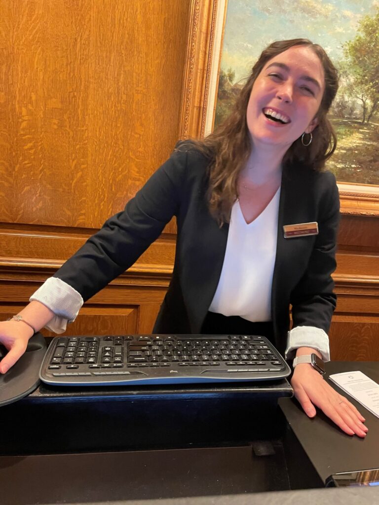 A woman, Mackenzie Mathews, at front desk of Biltmore Inn. She is wearing a white blouse with a dark jacket and there is a computer keyboard in front of her