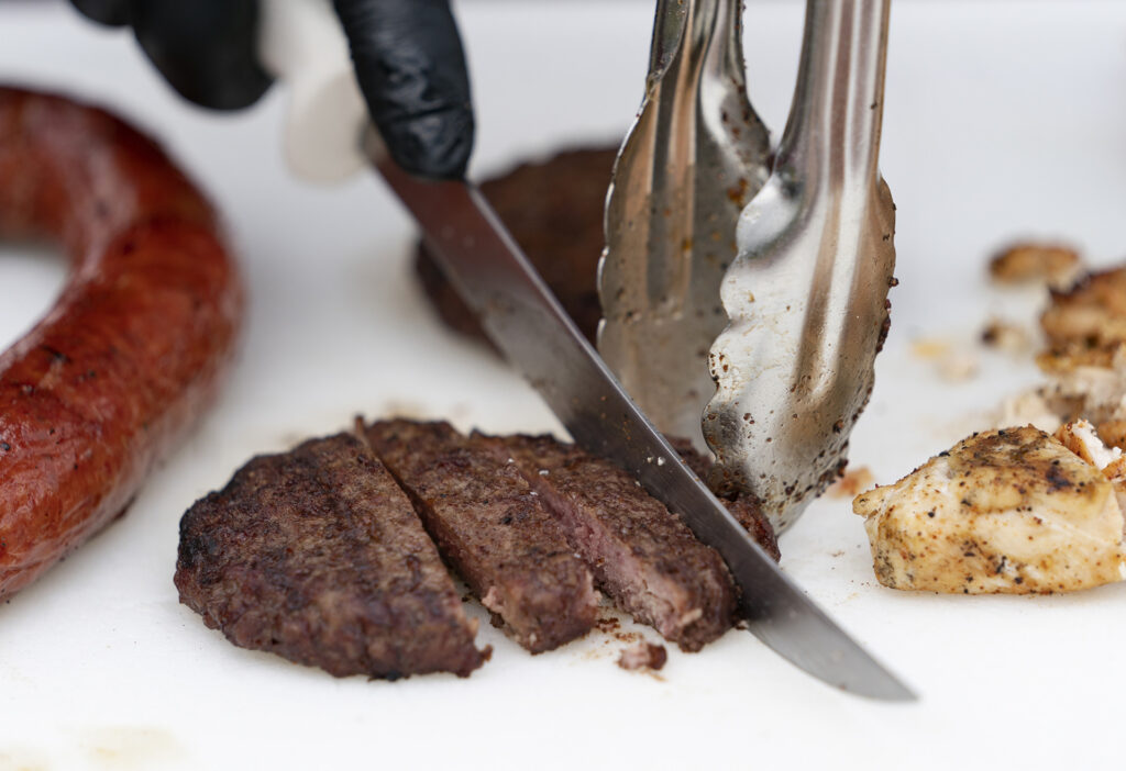 a picture of a hamburger patty being cut with a knife while held with tongs. There are pieces of chicken and sausage to the sides