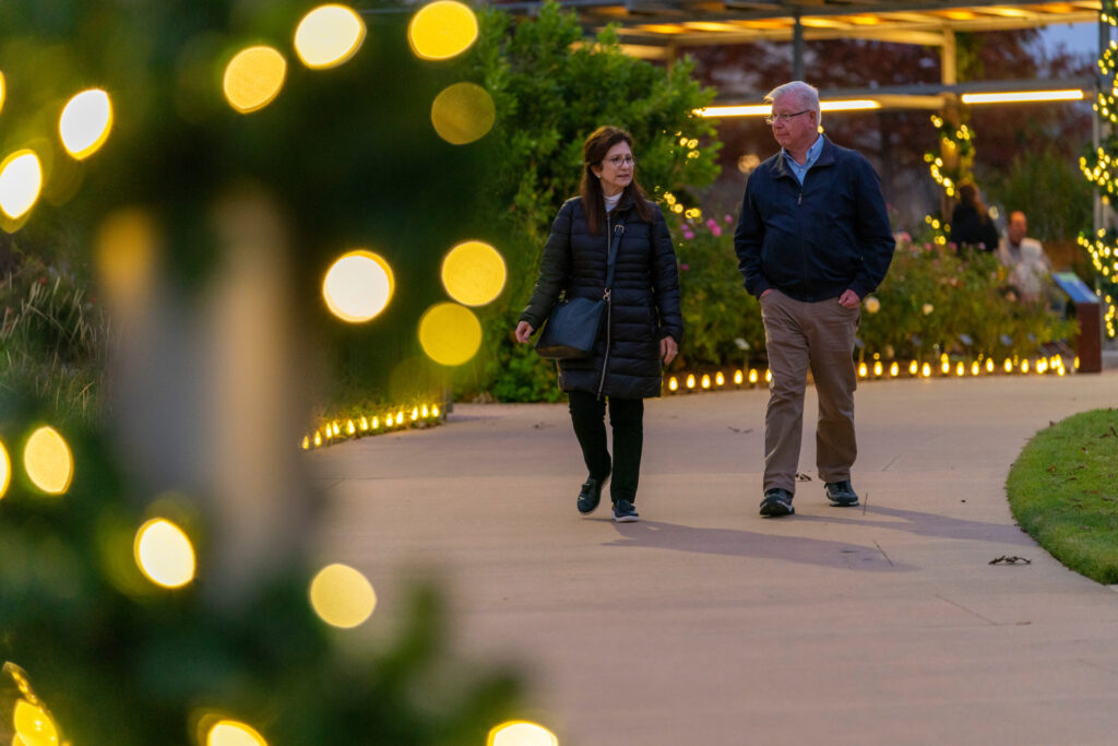 Middle-age couple stroll down sidewalk with lights in the foreground.