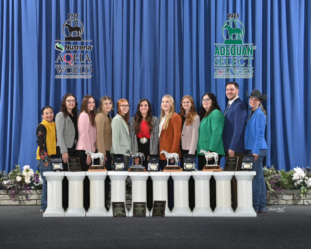 The Texas A&M horse judging team with 10 women and one man line up behind a row of horse statues and plaques with signs behind them indicating it is the AQHA World Championship.