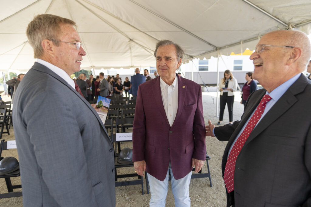 Three men speaking with each other during the groundbreaking ceremony. The man on the left is wearing a gray jacket, the man in the middle has a maroon jacket and the man on the right is wearing a dark jacket with a blue shirt and red tie. 