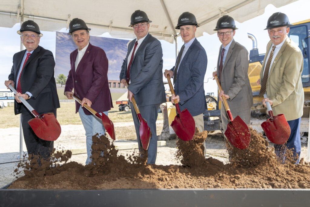 Five men with shovels turning dirt at a groundbreaking ceremony. They are all wearing suits and hard hats. 