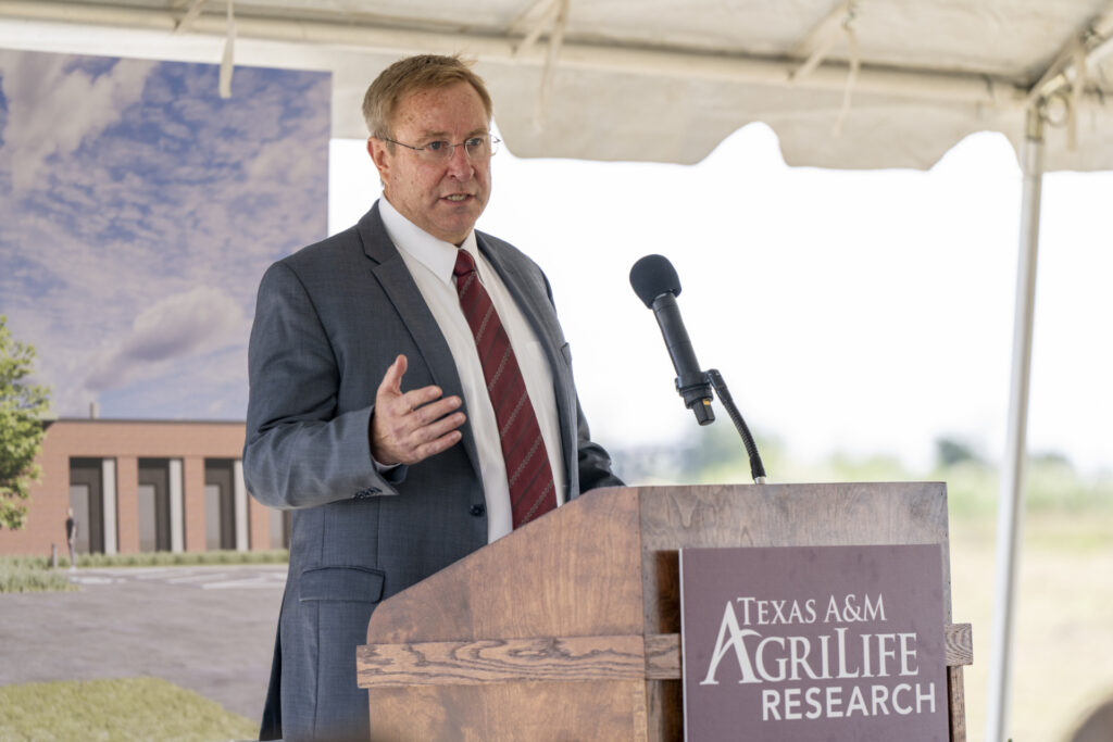 A man, G. Cliff Lamb, Ph.D., standing behind a podium speaking to a group of people. He is wearing a grey suit with a white shirt and maroon tie. The podium in front of him has a sign that says Texas A&M AgriLife Research.