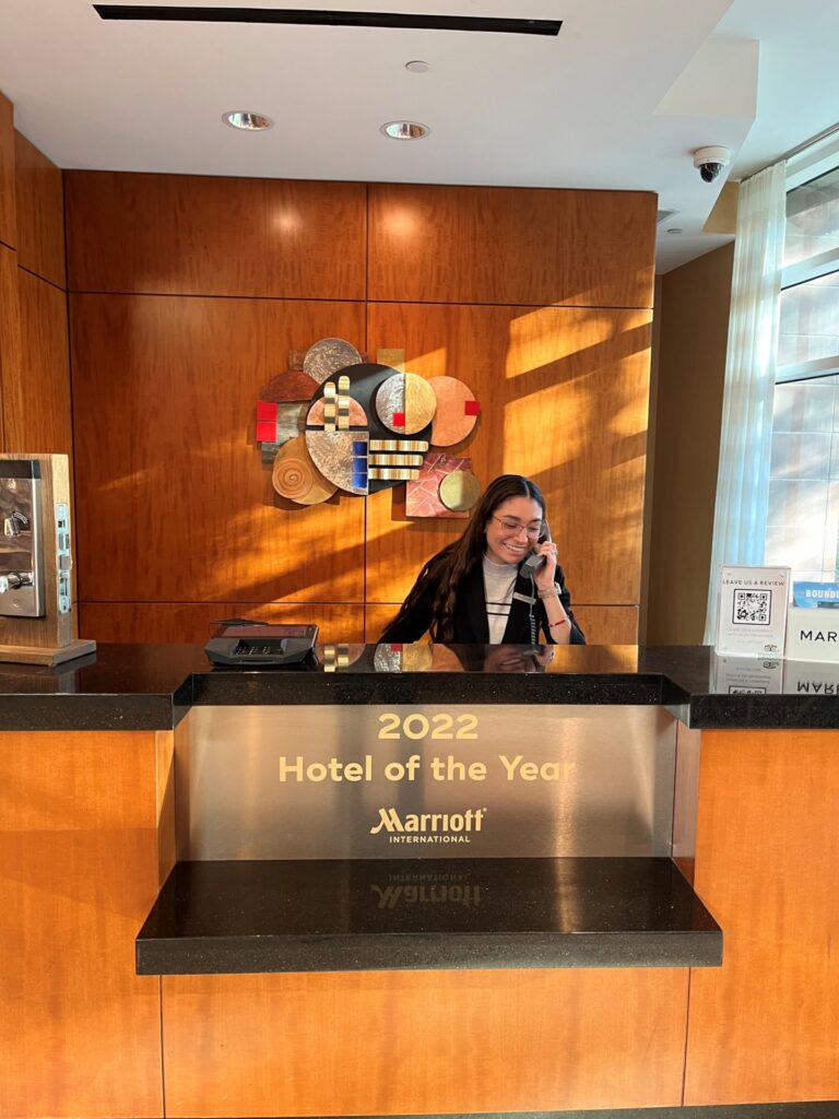 A woman, Yaadire Correa, sitting behind a reception desk in the lobby of The Woodlands Waterway Marriott Hotel and Convention Center. She is wearing a dark jacket and is on the phone. 