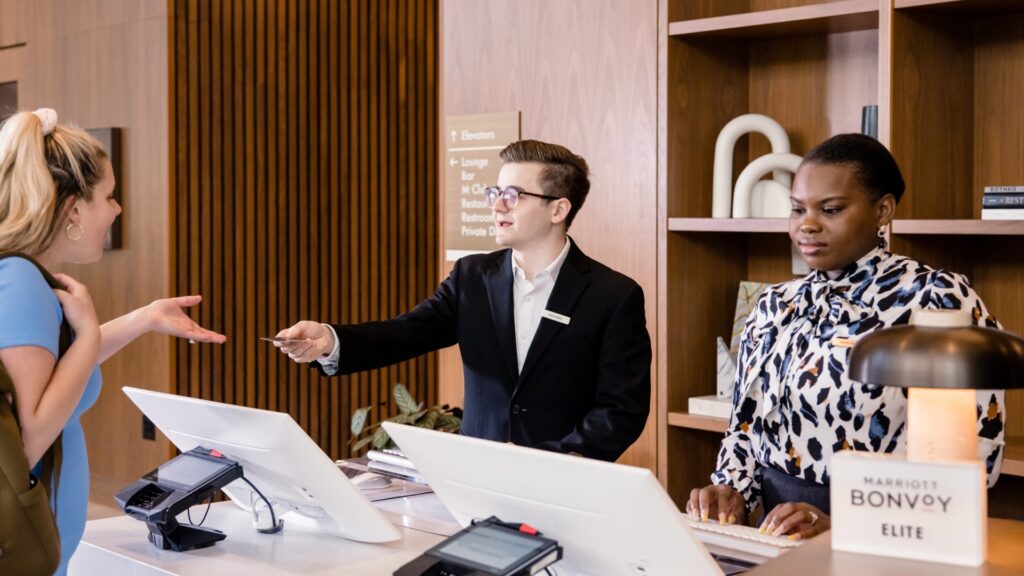 A women and a man behind a reception desk at a hotel with a woman checking in. The man is handing the guest her room key. 
