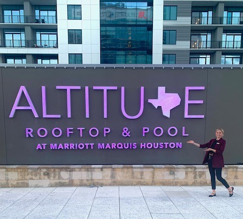 A woman dressed in a black jacket and pants, Shianne Holmes, in front of a sign that reads Altitude Rooftop & Pool at Marriott Marquis Houston. Holmes is part of the Voyage Program through Marriott.