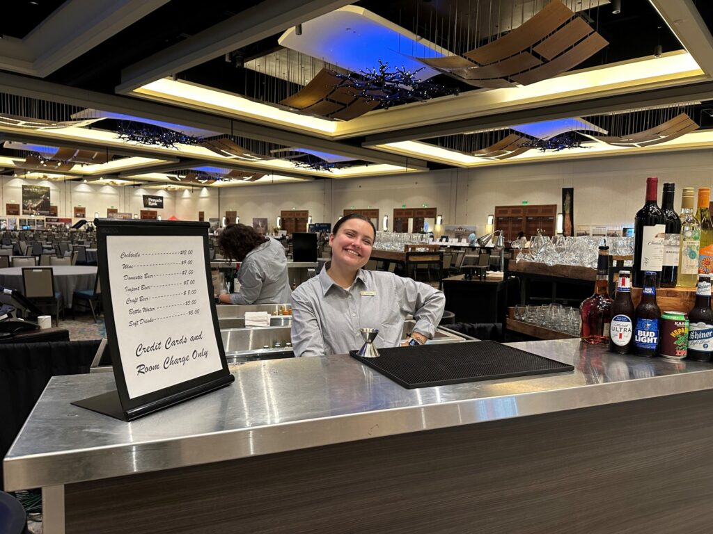 A woman, Taylor Stokes, standing behind the bar at the Marriott Hill Country Resort and Spa. She is wearing a blue short and there are bottles and a sing on the counter in front of her.  