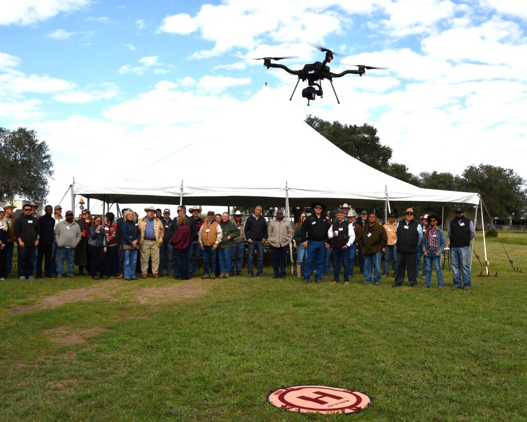 UAV in air with attendees of the Beeville Field day standing under a tent watching. 