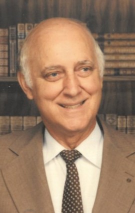 Head and shoulder photo of Leslie Reid, Ph.D. He is wearing a brown jacket with a white shirt and a brown tie with white dots on it. The backdrop is a canvas with books on it. 