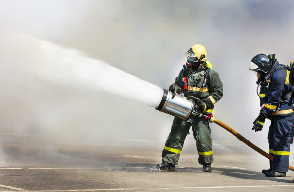 Two fully dressed out firefighters hold a hose with a wide nozzle that is spraying foam onto a fire. The foam contains PFAS, also known as forever chemicals.