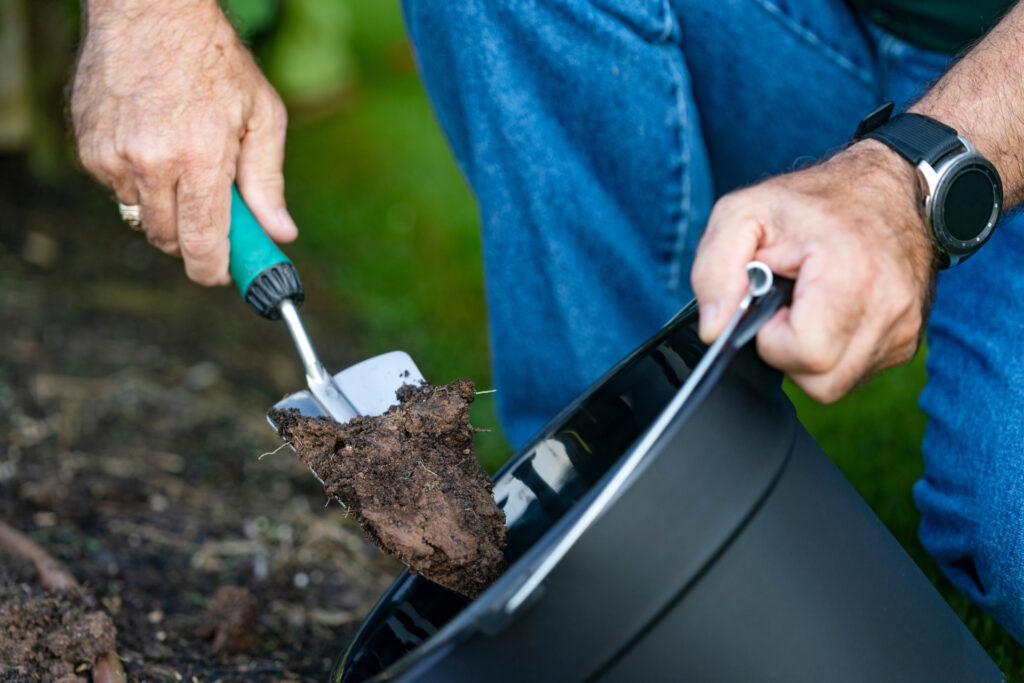 A man's hands hold a pot and a trowel in a gardeb. He is filling the pot with soil.